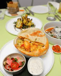 A dish of prawn biryani from COMO Cuisine, served in a pot with its dough lid broken open, accompanied by sides of raita and a pickled onion and tomato salad.