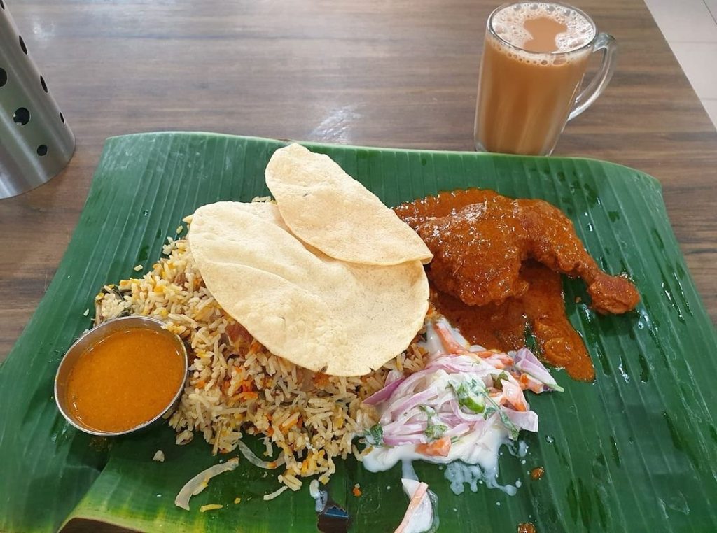 Indian meal from ASHMI Restaurant served on a fresh banana leaf, featuring chicken biryani with a whole chicken leg quarter in rich gravy, a side of creamy vegetable salad, papadums, and a small metal bowl of curry sauce.