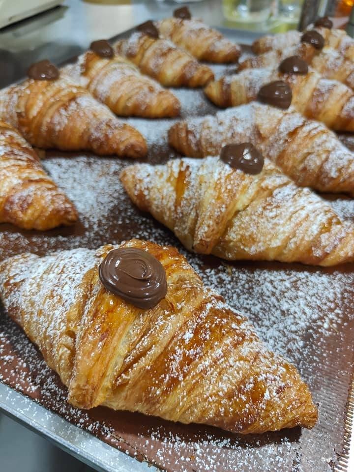 A tray of freshly baked golden croissants from Brunetti, each topped with a dollop of chocolate spread and generously dusted with powdered sugar.