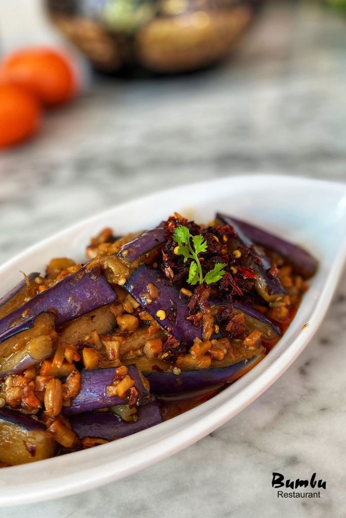 A dish of spicy stir-fried eggplant from Bumbu Restaurant, featuring sliced purple eggplant in a savory sauce with minced ingredients and chili flakes, garnished with cilantro, served in a white oval dish on a marble tabletop.