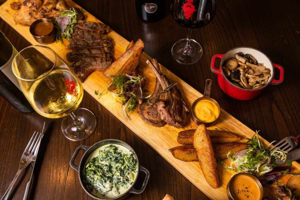 A lavish meat platter from Ginett Restaurant & Wine Bar, showcasing grilled chicken, steak, lamb chops, potato wedges, and sauces, served with sides of creamed spinach, sautéed mushrooms