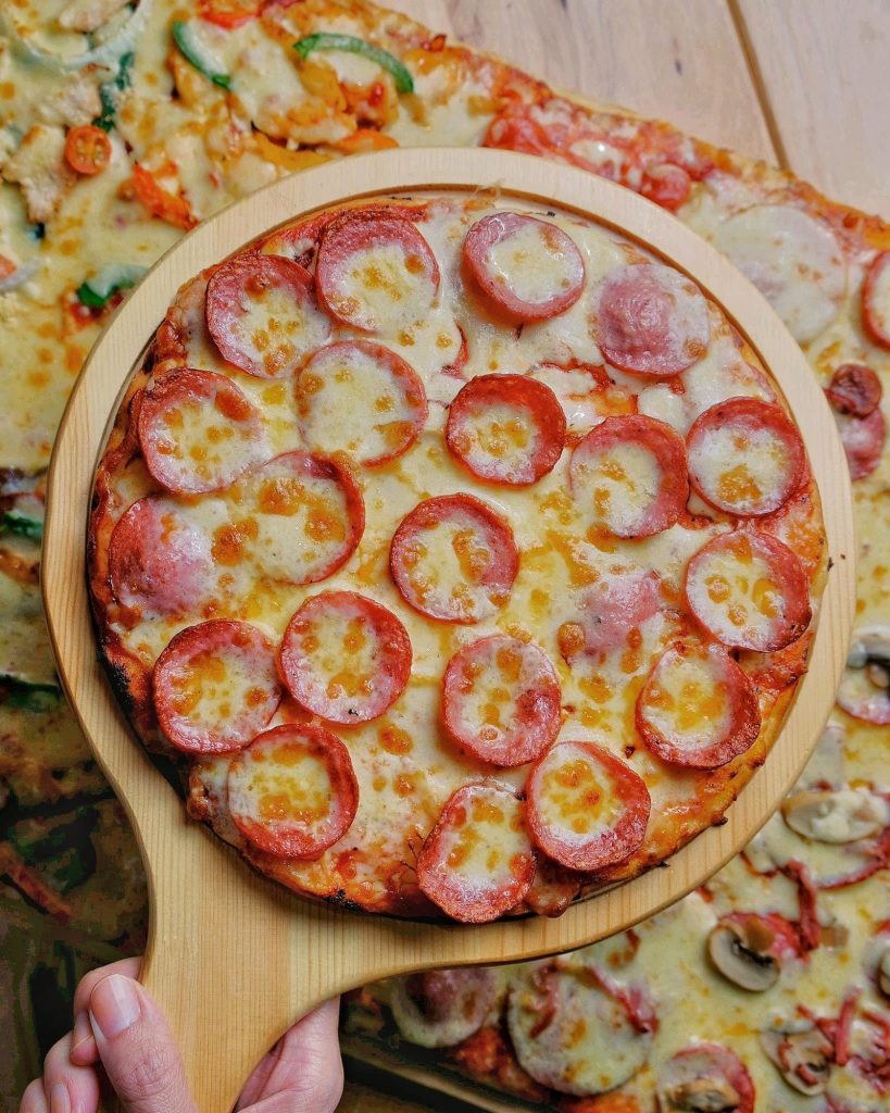 A hand holding a wooden pizza peel with a freshly baked pepperoni pizza