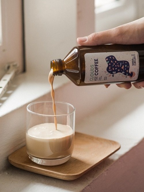 A hand pouring 'Clouds Coffee' cold brew from Grids & Circles from an amber glass bottle with a distinctive label into a clear glass