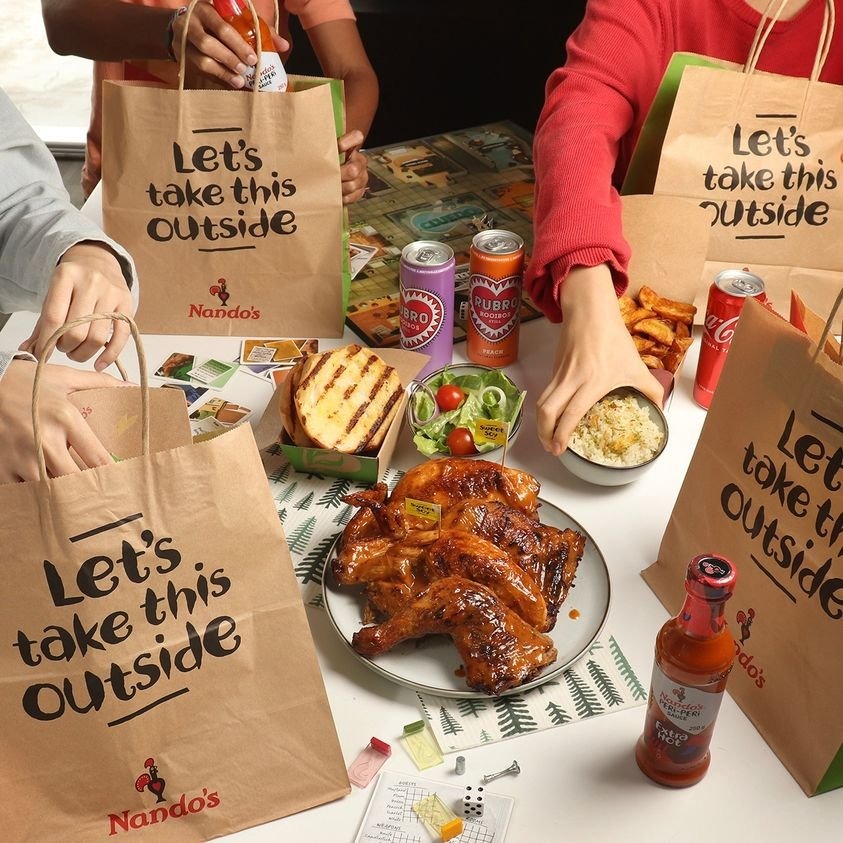 A Nando's takeaway spread from The Star Vista with a whole Peri-Peri chicken, sides, drinks, and Nando's sauce, surrounded by Nando's bags encouraging outdoor dining, with a board game indicating a social event.