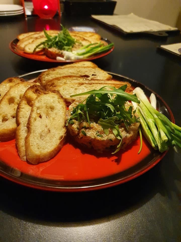 Two red plates on a dark table, each with a serving of tartare topped with arugula, alongside toasted bread slices and spring onions