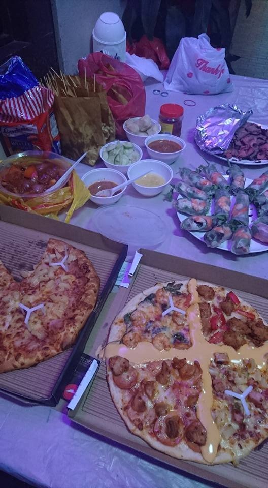 A table laden with a casual party feast: two open pizza boxes (one Hawaiian, one four-flavor with cheese sauce), a platter of fresh Vietnamese spring rolls, various dips, a saucy dish, and snack bags