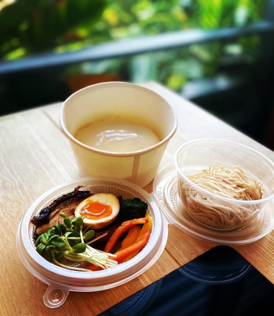Ramen takeaway set with separate containers for broth, noodles, and toppings (egg, pork, vegetables) on a wooden table