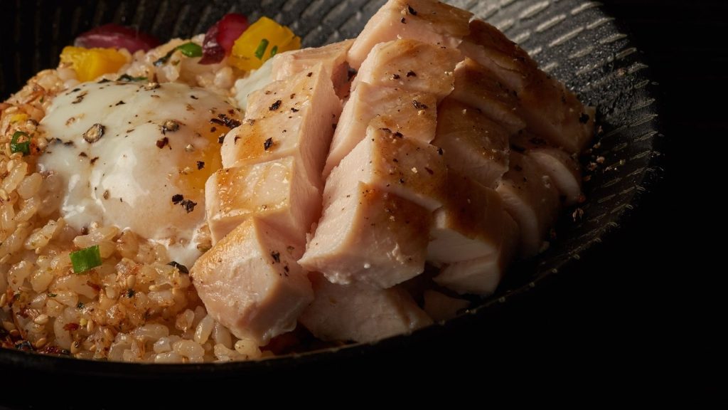 Japanese rice bowl with grilled chicken, soft-poached egg, and seasoned rice in a dark bowl.