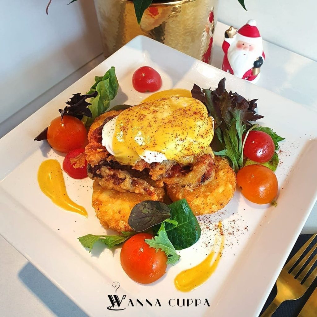 Wanna Cuppa's signature Eggs Benedict served on hash browns with a crispy fried topping, hollandaise sauce, fresh cherry tomatoes, and mixed greens, garnished with sauce swirls.