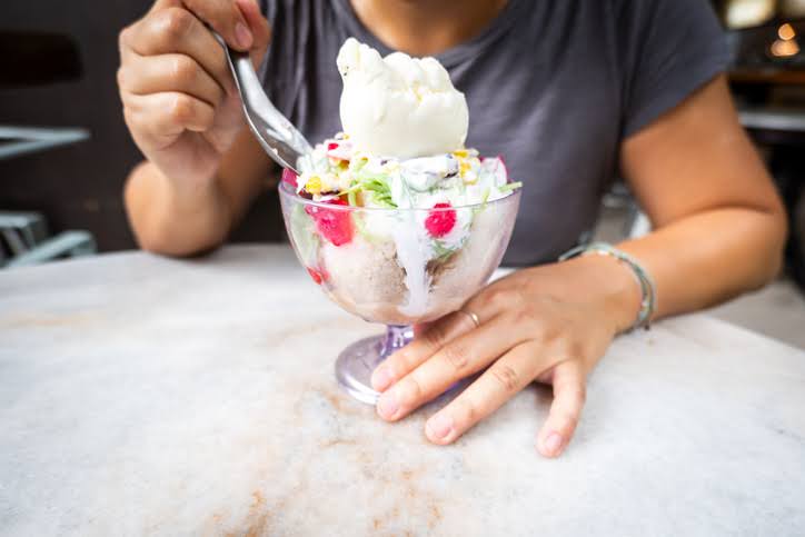 A person sits at a marble table, holding a spoon over a large glass of ice kacang